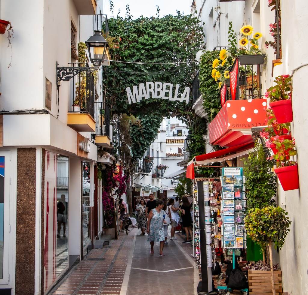 a picture of a street lined with flowers and a white sign strung up in the middle which says marbella